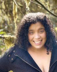 Image: Maxine Wright, a Black, light-skinned woman with curly hair, stands in the foreground smiling into the camera, with mossy trees as the backdrop